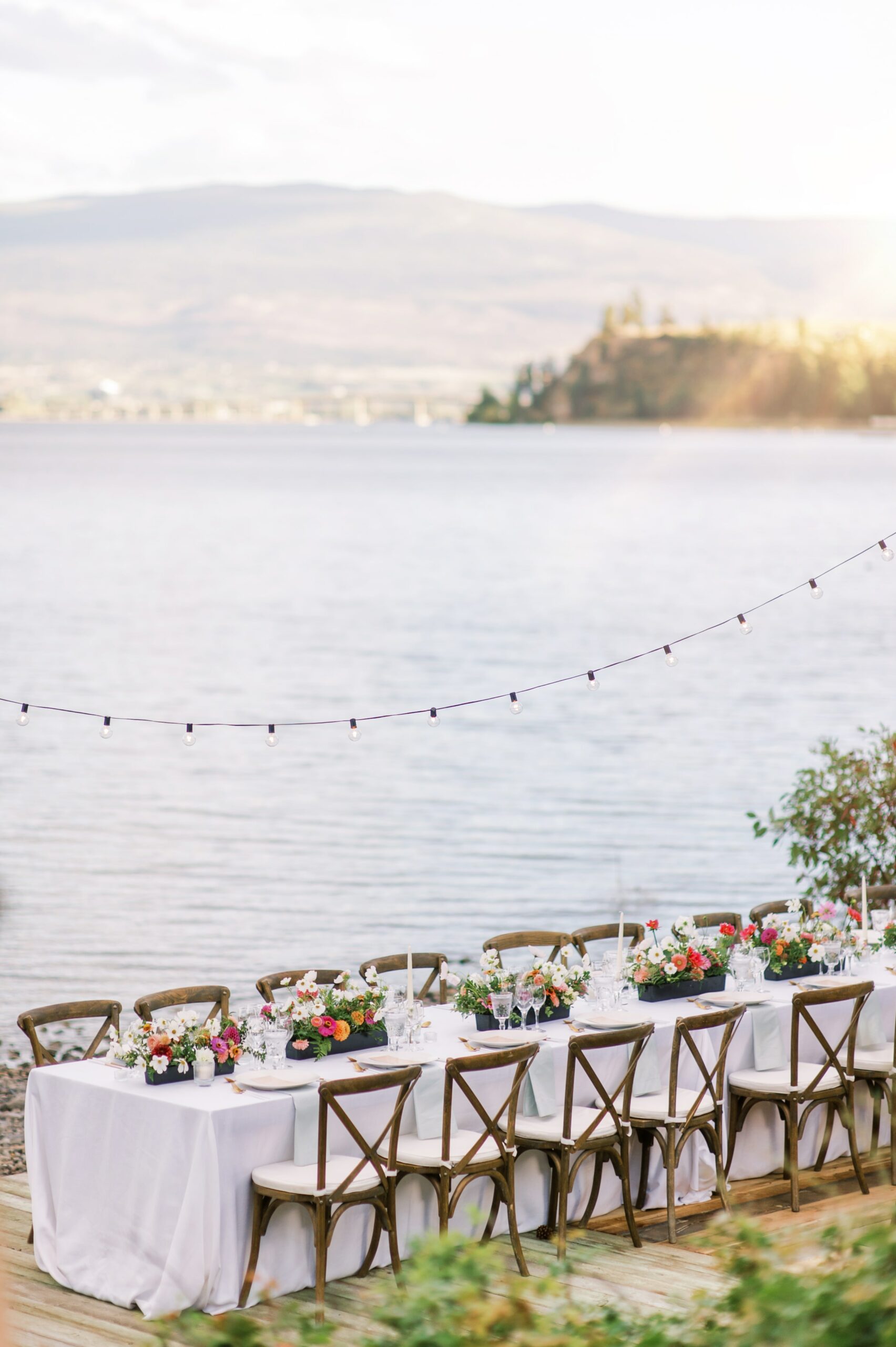 Long table dinner with white linens, wood chairs and colourful flowers in front of a lake